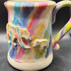 Puede incluir: Taza de cerámica hecha a mano con un esmalte colorido y arremolinado en tonos de azul, rosa, amarillo y verde. La taza presenta un diseño de oso esculpido en el lateral y un asa a juego. La taza mide aproximadamente 10 cm de alto.