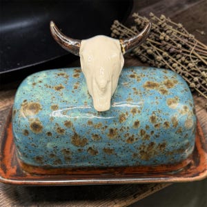 May include: A ceramic butter dish with a rectangular base and a domed lid. The lid features a cream-colored bull skull handle with dark brown horns. The lid has a mottled blue and brown glaze, while the base has a solid dark brown rim.