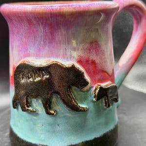 May include: A ceramic mug with a bear family design. The mug features a brown bear with the word "MAMA" and a smaller bear cub. The mug has a colorful glaze with pink, blue, and red hues.