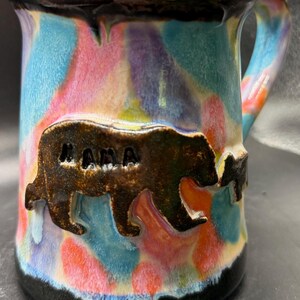 Puede incluir: Taza de cerámica hecha a mano con un diseño abstracto colorido en tonos de azul, rosa, morado y naranja. La taza presenta un diseño de oso pardo y osezno con la palabra "MAMA" tallada en el oso. El borde y la base son negros.