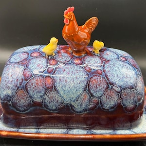 May include: A ceramic butter dish with a blue and red mottled pattern. The lid is topped with a brown rooster and two yellow chicks. The dish sits on a matching rectangular base.