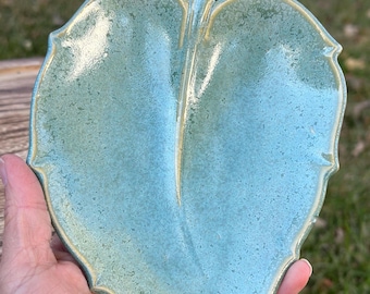 May include: A ceramic leaf-shaped dish with a light blue and green glaze. The dish has a textured surface and a slightly raised vein running down the center.