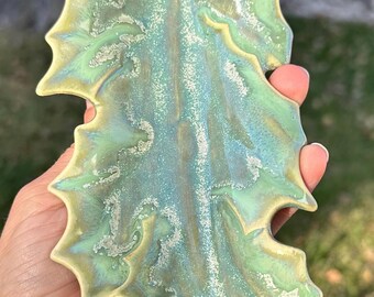 May include: A ceramic leaf dish with a green and blue glaze. The dish has a textured surface and a slightly iridescent finish.