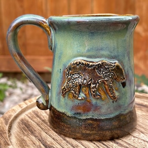 Puede incluir: Una taza de cerámica verde con un diseño de oso marrón. La taza tiene un esmalte marrón y un borde marrón oscuro.