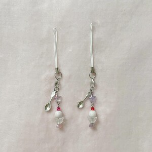 Op de afbeelding: Twee handgemaakte telefoonbedels met witte koorden, zilverkleurige metalen sluitingen en decoratieve bedels. Elke bedel heeft een kleine lepel, roze en paarse kralen en een wit, engelachtig kralenontwerp.