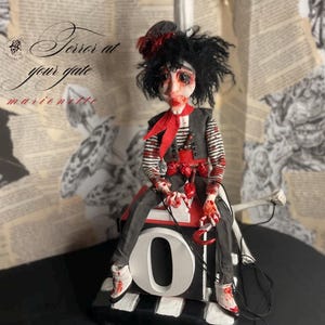 May include: A marionette doll with messy black hair, wearing a striped shirt, dark vest, and red neckerchief, sits on a block marked with the letter O. The doll features red paint splatters, a small hat, and holds a hook. Text reads Terror at your gate.