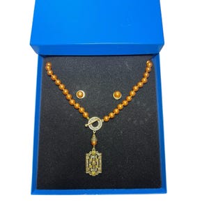 Könnte beinhalten: Ein Halsketten- und Ohrring-Set in einer blauen Geschenkbox. Die Halskette hat bernsteinfarbene Perlen, einen dekorativen Verschluss und einen rechteckigen Anhänger. Die passenden Ohrringe sind runde Ohrstecker. Das Set wird auf schwarzem Samt präsentiert.