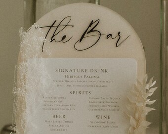 Op de afbeelding: Een wit bord met een zwarte boog bovenaan waarop in cursief "the Bar" staat. Het bord vermeldt een signature drankje, "Hibiscus Paloma", en een selectie van sterke drank, bier en wijn.