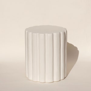 May include: White cylindrical stool with a fluted design. The stool is made of a smooth, white material and has a rounded top.