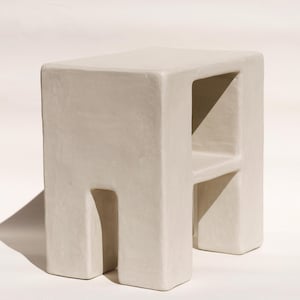 May include: A neutral-toned, square stool with a minimalist design. The stool features a solid top and base, with cut-out sections forming the legs and a shelf. The overall aesthetic is modern and sculptural, suitable for various interior styles.