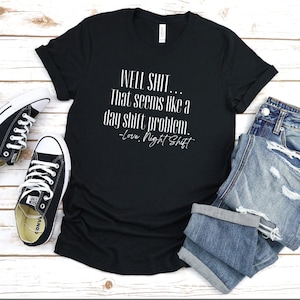 May include: Black t-shirt with white text that reads "WELL SHIT... That seems like a day shift problem. -Love, Night Shift". The shirt is laid out on a wooden surface with a pair of black converse shoes and a pair of blue jeans.