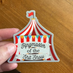 May include: A sticker featuring a circus tent design with red and white stripes, a red flag, and a banner that reads "Ringmaster of the Shit Show". The sticker has a gray border and is held against a wooden surface.