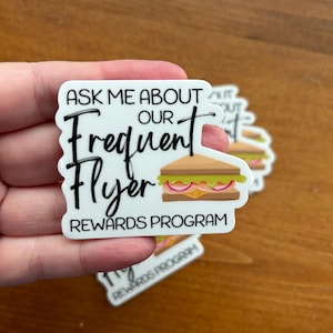 K&ouml;nnte beinhalten: Wei&szlig;er Aufkleber mit schwarzem Text, der "Ask me about our Frequent Flyer Rewards Program" lautet. Neben dem Text ist ein Cartoon-Sandwich abgebildet.