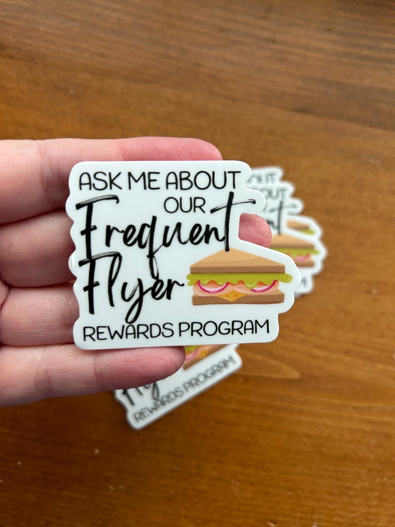 K&ouml;nnte beinhalten: Wei&szlig;er Aufkleber mit schwarzem Text, der "Ask me about our Frequent Flyer Rewards Program" lautet. Neben dem Text ist ein Cartoon-Sandwich abgebildet.