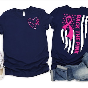 May include: Two navy blue t-shirts with a pink ribbon and stethoscope design. The front shirt has a pink ribbon and stethoscope in a heart shape. The back shirt has a pink ribbon on a white and blue American flag design with the text "Back the Pink".