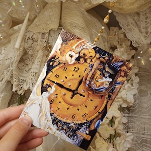 May include: A rectangular postcard featuring a surrealist clock design with an owl and angel figures. The clock face is gold with black numerals. The postcard is held against a backdrop of lace, flowers, and decorative beads.