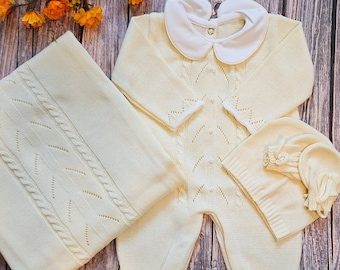 May include: A cream-colored knitted baby outfit with a white collar, matching hat, and a blanket. The outfit has a delicate cable knit pattern.