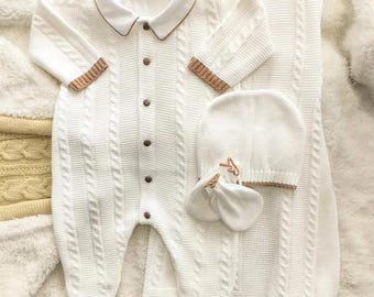 May include: A white cable-knit baby outfit set, including a footed onesie, hat, mittens, and blanket. The set features brown trim on the collar, cuffs, and accessories. The onesie has brown buttons down the front.