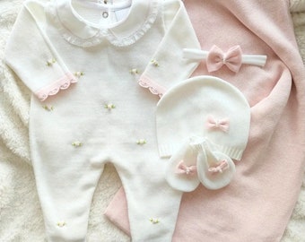 Può includere: Un completo per neonato in maglia bianca con bordini rosa e fiori ricamati, un berretto in maglia bianca con un fiocco rosa e un paio di scarpine in maglia bianca con fiocchi rosa.