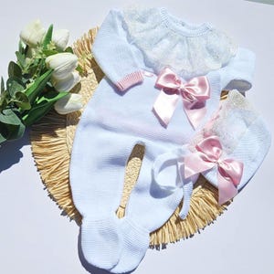 Könnte beinhalten: Ein weißer, gestrickter Strampler mit Füßen, langen Ärmeln, Spitzenkragen und einer rosa Satinschleife auf der Brust. Eine passende weiße Strickmütze mit Spitzenbesatz und rosa Satinschleife ist enthalten. Das Set hat hellrosa gerippte Ärmelbündchen.