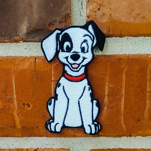 May include: A white and black embroidered patch of a cartoon Dalmatian dog sitting with a red collar. The dog has black spots and a happy expression.