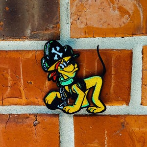 Op de afbeelding: Een gele en zwarte cartoon hondenpatch met een piratenhoed met een schedel- en gekruiste botten ontwerp. De hond heeft een groene kraag en een rode tong. De patch zit op een bakstenen muur.