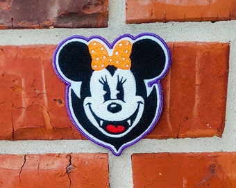 May include: A black and white embroidered patch of a cartoon mouse with fangs, wearing a purple and orange bow. The mouse is smiling with its fangs showing.