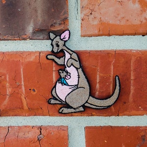 May include: A brown and gray embroidered patch of a kangaroo carrying a joey in its pouch. The kangaroo is standing on its hind legs with its tail curled around its body. The joey is peeking out of the pouch.