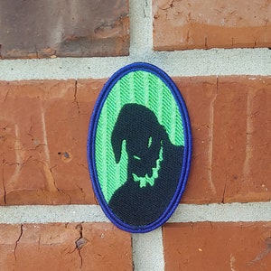Op de afbeelding: Een geborduurde patch met een silhouet van Oogie Boogie uit de film The Nightmare Before Christmas, in groen en blauw.