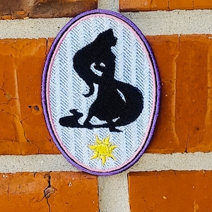 Op de afbeelding: Een ovaal gestikt patch in paars en wit met een silhouet van een vrouw met lang haar en een zon met gele stralen.