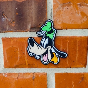 May include: A cartoon character patch of Goofy with a green hat, brown fur, and a yellow nose, stitched onto a brick wall.