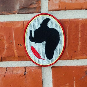 以下が含まれることがあります： 赤い、白、黄色の刺繍が施されたパッチで、くちばしに赤い羽根を持った黒い鳥のシルエットが描かれています。パッチはレンガの壁にあります。