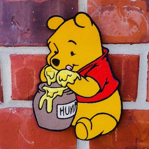 Könnte beinhalten: Ein gestickter Winnie Puuh-Charakter-Patch, gelb mit einem roten Hemd, der einen braunen Honigtopf mit gelbem Honig hält, der von oben tropft. Der Patch befindet sich an einer roten Backsteinwand.