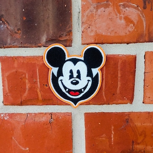 Op de afbeelding: Een geborduurde patch van Mickey Mouse met een ondeugende grijns en tanden. De patch is zwart-wit met een oranje rand en is bevestigd aan een bakstenen muur.
