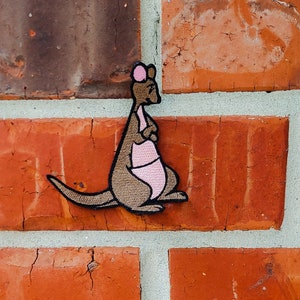 May include: A brown kangaroo with pink ears and a pink pouch is embroidered on a brick wall.