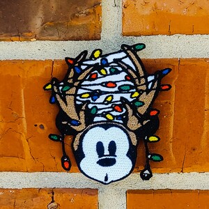 Op de afbeelding: Geborduurde patch met Mickey Mouse met geweien gemaakt van kerstverlichting. De patch heeft een wit gezicht, zwarte oren en bruine geweien versierd met kleurrijke lampjes. De patch is geplaatst tegen een bakstenen muur.