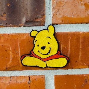 Op de afbeelding: Geborduurde patch met een lachende gele beer met een rood shirt. De patch is bevestigd aan een rode bakstenen muur. De beer heeft zwarte ogen en een zwarte neus. De patch is een stripfiguur.