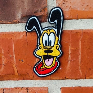 Op de afbeelding: Een geborduurde patch met een cartoonhond met een geel gezicht, zwarte oren en een rode halsband. De hond heeft een brede glimlach en steekt zijn tong uit.