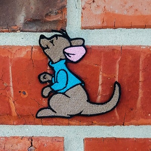 以下が含まれることがあります： 青いシャツを着てピンクの耳をした、漫画のカンガルーの刺繍パッチ。カンガルーは上を見て右を向いています。