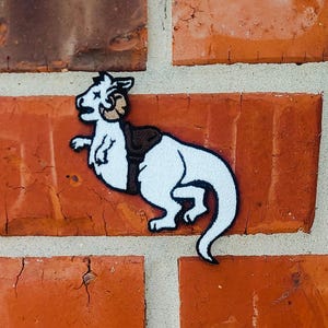 May include: White embroidered patch of a creature with a saddle on its back. The creature has a large head with horns and a long tail.