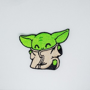 Puede incluir: Parche bordado verde y beige de un personaje de dibujos animados con ojos grandes y una sonrisa, que lleva una túnica. El personaje es conocido como "Baby Yoda" de la serie Star Wars.