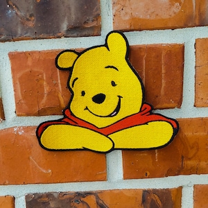 May include: A yellow embroidered Winnie the Pooh patch with a red shirt, smiling and looking to the right. The patch is attached to a brick wall.