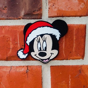 Op de afbeelding: Een rood en wit kerst-geborduurd patch met een cartoonfiguur met een rode kerstmanmuts, zwarte oren en een wit gezicht. Het personage heeft zwarte ogen, een zwarte neus en een roze mond.