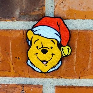 May include: A yellow Winnie the Pooh character wearing a red Santa hat with a white trim and a jingle bell. The character is smiling and has a black nose.