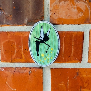 Peut inclure: Un patch brodé vert et blanc représentant la fée Clochette, un personnage de Peter Pan de Disney, avec une baguette magique et de la poussière de fée. Le patch est de forme ovale et a une bordure blanche.