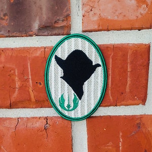 May include: A green and white oval-shaped patch with a black silhouette of Yoda's head and the Star Wars logo in green.
