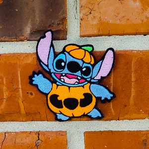 Könnte beinhalten: Ein gestickter Aufnäher von Stitch aus Lilo & Stitch, der sich für Halloween als Kürbis verkleidet hat. Der Aufnäher ist blau und orange mit schwarzem Umriss und das Wort "BOO" ist auf dem Kürbis zu sehen.