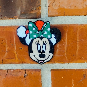 May include: An embroidered patch of Minnie Mouse wearing a Santa hat with white trim and a green bow with white polka dots.