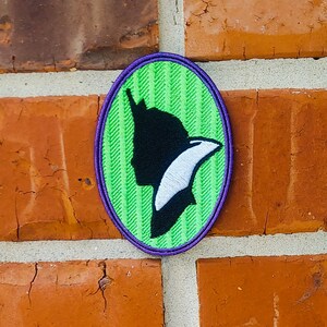 May include: A green and purple oval-shaped patch with a black silhouette of a person wearing a hat and a white bird flying above them.