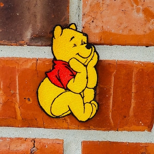 Può includere: Un toppa ricamata di Winnie the Pooh di colore giallo con una camicia rossa, seduto con le mani giunte sotto il mento, guardando in alto e a destra. La toppa è attaccata a un muro di mattoni.